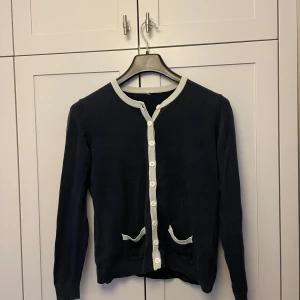 Svart kofta med vita detaljer - Snygg marinblå kofta med vita knappar och kantdetaljer. Den har långa ärmar och två små fickor fram.