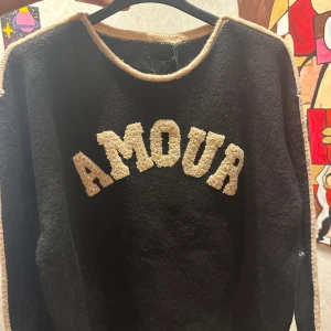 Svart tröja med texten 'AMOUR' - Säljer en svart tröja med beige detaljer och texten 'AMOUR' i pärlor på framsidan. dem för 500-550 lr då dem är ny! Tröjan har långa ärmar och är perfekt för en mysig höstlook. Den är i bra skick och passar både till vardags och lite finare tillfällen. 