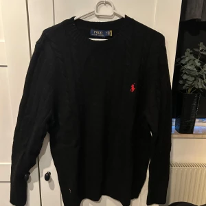 Svart tröja från Polo Ralph Lauren - Säljer en klassisk svart tröja från Polo Ralph Lauren i storlek M. Tröjan har en röd broderad logga på bröstet och är i mycket bra skick. Perfekt för både höst och vinter, med långa ärmar och en stilren design.