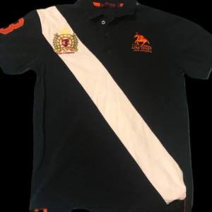 Svart och vit polo💯 - Snygg svart polo med en vit diagonal rand och broderad logga på bröstet. Den har korta ärmar och en klassisk krage med knappar. Perfekt för en avslappnad men stilren look. Passar både till vardags och lite finare tillfällen.