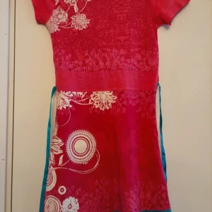 Röd mönstrad klänning från Desigual - Säljer en färgglad röd, unik klänning från Desigual med unikt blommönster och turkost knytband i midjan. Klänningen har korta ärmar och är perfekt för sommarens fester. Materialet känns mjukt och bekvämt, vilket gör den idealisk för både vardag och speciella tillfällen. 🌺Köparen betalar frakten. 