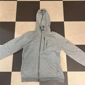 Zip up hoodie från Sail Racing - Säljer en grå zip up hoodie från Sail Racing i  bra skick. Den har en dragkedja framtill och en praktisk ficka med dragkedja på bröstet. Perfekt för kyliga dagar och passar till det mesta. Huvan är skön och ger extra värme. Perfekt för både vardag och fritid! Priset kan diskuteras vid snabb affär