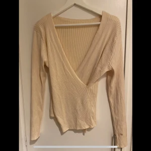 Beige ribbad omlottopp - Säljer en superfin beige ribbad omlottopp med v-ringning. Den är långärmad och perfekt för både vardag och fest. Materialet är mjukt och stretchigt, vilket gör den bekväm att bära hela dagen. Passar perfekt till jeans eller kjol!