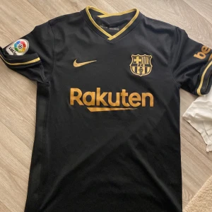 Svart FC Barcelona fotbollströja - Säljer en svart FC Barcelona tröja från Nike i mycket bra skick. Den har korta ärmar och snygga guldiga detaljer med klubbens emblem och Rakuten-logga på framsidan. La Liga-märket sitter på ärmen. Perfekt för alla Barça-fans! ⚽️