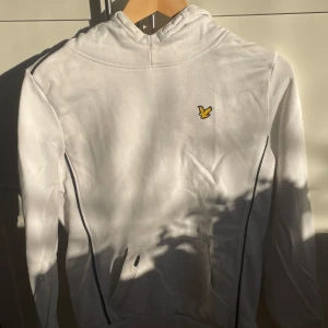 Vit hoodie från Lyle & Scott - Säljer en stilren vit hoodie från Lyle & Scott i mycket bra skick. Den har en klassisk design med en gul logga på bröstet och svarta detaljer längs sidorna. Perfekt för både vardag och chill. Passar bra till jeans!