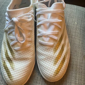 Vita och guldiga fotbollsskor från Adidas - Säljer ett par snygga vita fotbollsskor från Adidas med guldiga detaljer. Skorna har en modern design med snörning och är i mycket bra skick. Perfekta för fotbollsträning eller match. Sulan är designad för bra grepp på planen.