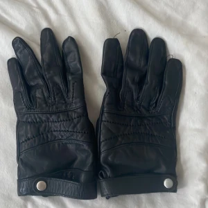 Svarta skinnhandskar med knäppning - Säljer ett par snygga svarta skinnhandskar i damstorlek. De har en stilren design med knäppning vid handleden för extra stil och passform. Perfekta för vintern och håller händerna varma och skyddade. Passar både till vardags och mer formella tillfällen.Storlek S/M