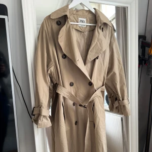 Beige trenchcoat från Zara - Säljer en klassisk beige trenchcoat från Zara i mycket bra skick. Den har långa ärmar och ett bälte i midjan för en snygg passform. Perfekt för höst och vår! 🧥Nypris 1200. Använda 1 gång.