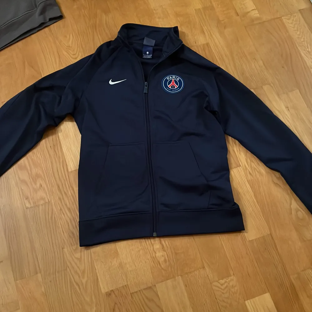 Säljer en snygg mörkblå träningsjacka från Nike med PSG-logga på bröstet. Jackan har dragkedja och långa ärmar, perfekt för fotbollsträning eller vardagsbruk. Den är i mycket bra skick och passar perfekt för höst och vår.. Jackor.