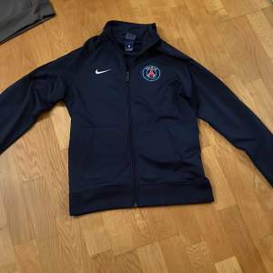 Säljer en snygg mörkblå träningsjacka från Nike med PSG-logga på bröstet. Jackan har dragkedja och långa ärmar, perfekt för fotbollsträning eller vardagsbruk. Den är i mycket bra skick och passar perfekt för höst och vår.