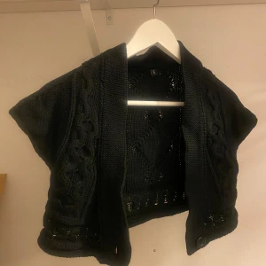 Svart stickad kofta från b.young - Säljer en snygg svart stickad kofta från b.young. Den har ett fint flätmönster och är perfekt för lager-på-lager under höst och vinter. Koftan är kortärmad och har en öppen design som gör den lätt att matcha med olika outfits.
