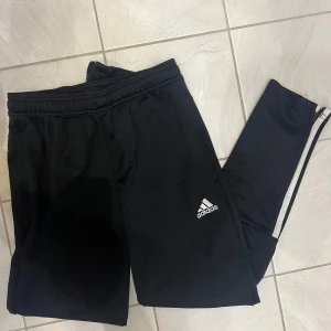 Svarta träningsbyxor från Adidas❤️ - Säljer ett par svarta träningsbyxor från Adidas i storlek 152. Byxorna har en elastisk midja och de klassiska tre ränderna längs sidorna. Perfekta för träning eller en avslappnad dag hemma. De är i mycket bra skick och gjorda av ett bekvämt syntetmaterial.