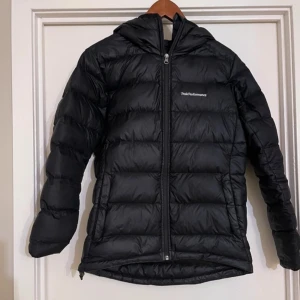 Svart dunjacka från Peak Performance - Säljer en svart dunjacka från Peak Performance. Den är perfekt för kalla dagar med sin varma vaddering och huva. Jackan har en dragkedja framtill och långa ärmar. Det finns en liten reva bak vid ryggslutet (har satt en bit tejp över men det går att ta bort och laga) men annars i jättebra skick. Perfekt för höst och vinter!