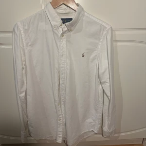 Vit skjorta från Ralph Lauren - Säljer en klassisk vit skjorta från Ralph Lauren i mycket bra skick. Skjortan har långa ärmar och en broderad logga på bröstet. Perfekt för både vardag och fest! 👔