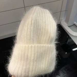 Vit stickad mössa - Säljer en supermysig vit stickad mössa i ribbat material. Perfekt för kalla vinterdagar och håller dig varm och stilren. Mössan är i mycket bra skick och passar de flesta tack vare sin stretchiga design. Ett måste för vintern! ❄️