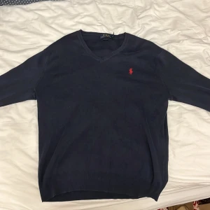 Mörkblå  v neck tröja från Polo Ralph Lauren - Säljer en snygg mörkblå tröja från Polo Ralph Lauren i Pima bomull. Den har en klassisk v-ringning och det ikoniska röda logotypbroderiet på bröstet. Perfekt för både vardag och lite finare tillfällen. Tröjan är i mycket bra skick och superbekväm att ha på sig.