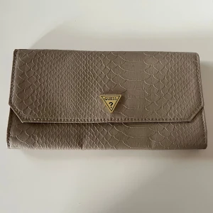 Beige plånbok från Guess - Snygg beige plånbok från Guess med ett coolt ormskinnsmönster. Den har en praktisk knappstängning och en transparent fönsterficka på insidan för ID eller kort. Perfekt för att hålla ordning på dina grejer med stil!