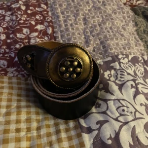 Brunt bälte med guld dekorativt spänne - Säljer ett snyggt brunt bälte med ett stort, dekorativt spänne i guld. Bältet har en unik design med nitar och detaljer som ger en vintagekänsla. Perfekt för att piffa upp en enkel outfit eller för en festlig look. Passar både till jeans och klänning!