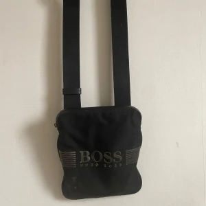 Svart axelväska från Hugo Boss - Snygg och stilren svart axelväska från Hugo Boss. Perfekt för att bära med sig det nödvändigaste. Väskan har en justerbar rem och ett diskret BOSS-tryck på framsidan. Passar både till vardags och festliga tillfällen.