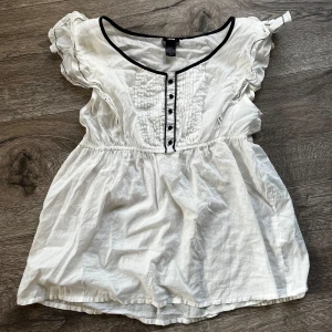 Vit blus med svarta detaljer - Söt vit blus från H&M med svarta knappar och kantdetaljer. Den har korta ärmar med volang och en v-ringad hals. Perfekt för vår och sommar, både till vardags och fest. Blusen är gjord i ett lätt och luftigt material som är något genomskinlig 💋