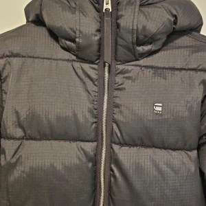 Svart pufferjacka från G-Star RAW - Säljer en svart pufferjacka från G-Star RAW i storlek S. Jackan har en dragkedja framtill och en mysig huva. Perfekt för kalla dagar och håller dig varm och stilren. Passar både till vardags och lite finare tillfällen. 🖤