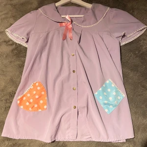 Lila kortärmad klänning med prickiga fickor - Supergullig lila klänning med korta ärmar och söta spetsdetaljer. Den har en peter-pan krage och en rosa rosett och knappar framtill. Det är en babydoll modell ger en A-linje siluett. Den har två färgglada fickor i rosa och blått med prickigt mönster ger en lekfull touch. 