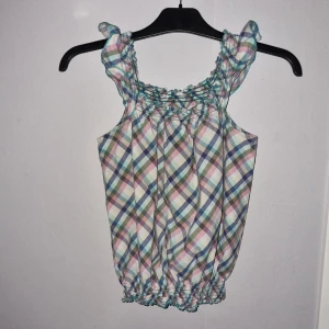 Rutig ärmlös topp - Söt ärmlös topp med rutigt mönster i pastellfärger. Den har volangdetaljer vid axlarna och en elastisk kant vid midjan.