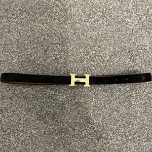 Svart skärp med guldfärgat spänne - Säljer ett elegant svart skärp i skinn imitation med ett stilrent guldfärgat H-spänne. Brun på insidan. Perfekt för att ge en lyxig touch till din outfit. Skärpet är i bra skick och passar både till vardag och fest. Ett måste för garderoben!