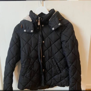 Svart quiltad jacka med huva - Säljer en snygg svart quiltad jacka med avtagbar huva och dragkedja. Perfekt för höst och vinter, håller dig varm och stilren. Jackan har långa ärmar och en mysig foder på insidan av huvan. Passar både till vardags och lite finare tillfällen.