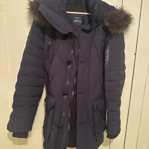 Mörkblå parkajacka från Superdry - Säljer en mörkblå parkajacka från Superdry i bra skick. Jackan har en mysig pälsfodrad huva och praktiska dragkedjor. Perfekt för kalla vinterdagar. Den är quiltad och har flera fickor för extra förvaring. Passar perfekt för både vardag och utflykter!