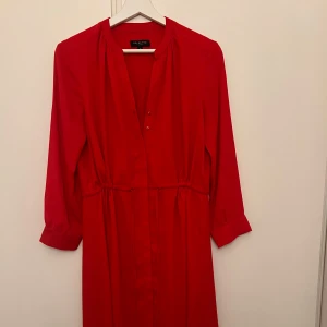 Röd klänning från Selected Femme - Snygg röd klänning från Selected Femme i storlek 36. Tillverkad av 100% återvunnen polyester, perfekt för både vardag och fest. Klänningen har långa ärmar och en stilren design med knappar framtill. Ett måste i garderoben för en färgklick!