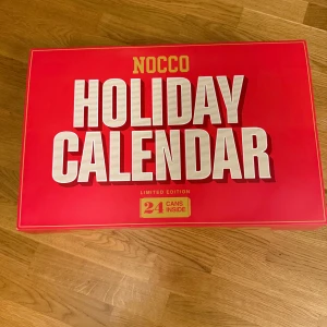 NOCCO Holiday Calendar - Säljer en NOCCO Holiday Calendar i en snygg röd färg. Denna limited edition-kalender innehåller 24 burkar, perfekt för att räkna ner till julen med en ny smak varje dag. Kalendern är stor och rektangulär, vilket gör den till en perfekt present eller för att unna sig själv något extra under högtiden.