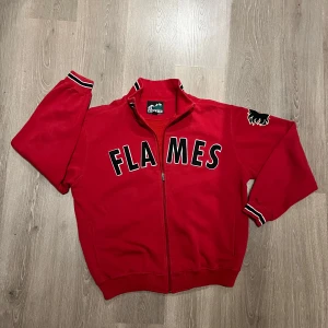 Röd Calgary Flames jacka/ tröja - Säljer en röd tröja/ jacka med Calgary Flames-logga och dragkedja framtill. Den har långa ärmar och ribbade muddar i svart och vitt. Perfekt för fans av Calgary Flames!