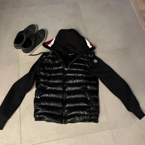 Moncler cardigan - Säljer en snygg svart jacka från Moncler med quiltad framsida och mjuka ärmar. Den har en dragkedja och en huva med röd och vit randdetalj.  Jackan är i mycket bra skick och redo för nya äventyr! missa inte detta. Jackan är väl omhändertagen och har inga som helst skråmor eller repor på glansen inga hål eller blekningar av något slag perfekt till sena sommar kvällar 