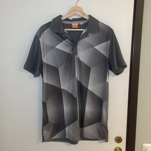 Grå mönstrad pikétröja från Puma - Säljer en snygg grå pikétröja från Puma med geometriskt mönster. Perfekt för sportiga aktiviteter eller en avslappnad dag. Tröjan har korta ärmar och en klassisk krage. Den är i mycket bra skick och redo för nya äventyr! Pris går alltid att förhandlas!