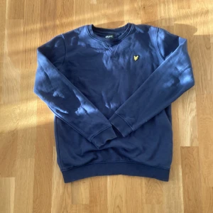 Blå tröja från Lyle & Scott - Säljer en snygg blå tröja från Lyle & Scott Junior. Den har en klassisk design med rund hals och ett litet gult emblem på bröstet. Perfekt för både vardag och skola. Tröjan är i bra skick och passar till alla säsonger.