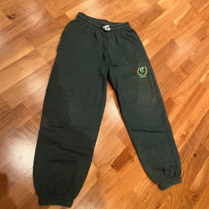 Gröna mjukisbyxor med Färjestad BK-logga - Säljer ett par gröna mjukisbyxor med Färjestad BK-logga. Byxorna har en bekväm resårmidja med snörning och muddar vid bensluten. Perfekta för en avslappnad dag hemma eller för att visa ditt stöd för laget. De är i bra skick och redo för nya äventyr!