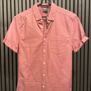 Rosa kortärmad skjorta från ASOS - Säljer en snygg rosa kortärmad skjorta från ASOS i storlek S. Skjortan är gjord av mjuk bomull med en liten andel elastan för extra komfort. Den har en klassisk krage och knappar framtill. Perfekt för sommardagar eller avslappnade tillfällen. 🌸