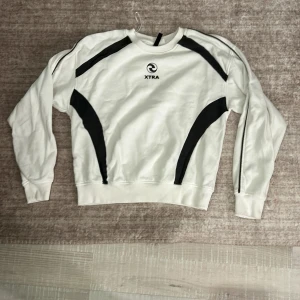 Vit och svart tröja från H&M Divided - Säljer en vit tröja från H&M Divided med svarta detaljer och XTRA-logga. Den är i bra skick och har långa ärmar. Perfekt för en avslappnad stil eller träning. Passar bra till både jeans och mjukisbyxor. Säljer för den blir aldig andvänd 