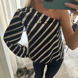 Randig oneshoulder topp - Säljer en superfin randig oneshoulder topp i svart och vit. Den har en unik design med en lång ärm och en axel bar, vilket gör den perfekt för både vardag och fest. Materialet känns mjukt och bekvämt mot huden. Passar perfekt med jeans eller kjol för en stilren look.