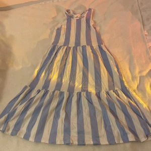 Randig sommarklänning - Säljer en superfin randig klänning i blått och vitt. Perfekt för sommardagar! Den är ärmlös och har en luftig passform. Klänningen är i bra skick och passar perfekt för en dag på stranden eller en sommarfest.