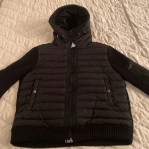 Moncler cardigan - Säljer en snygg svart pufferjacka med huva. Jackan har långa ärmar och dragkedja framtill. Perfekt för höst och vinter, håller dig varm och stilren. Den har ribbstickade muddar och två fickor med knappar. Passar både till vardags och lite finare tillfällen.