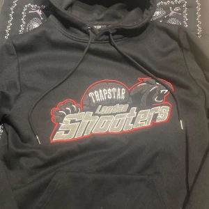 Svart trapstar hoodie - Säljer en svart hoodie från Trapstar med coolt tryck 'London Shooters' i grått och rött. Den har en klassisk känguruficka och justerbar huva med snören. Perfekt för en avslappnad stil eller streetwear-look. Passar bra till både jeans och joggers.