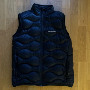 Navy dunväst från Peak Performance - Säljer en snygg navy dunväst från Peak Performance i mycket bra skick. Västen har en dragkedja framtill och är perfekt för höst och vinter. Den quiltade designen ger extra värme och stil. Passar både till vardags och lite mer uppklädda tillfällen. Är i princip nytt skick (har bara tagit bort lapparna). 