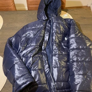 Mörkblå dunjacka från Ralph Lauren - Säljer en snygg mörkblå dunjacka från Ralph Lauren. Jackan har en glansig finish och är perfekt för kalla dagar. Den har en praktisk huva och dragkedja framtill. Perfekt för vintern och hösten!