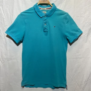 Turkos pikétröja från Hilfiger Denim - Säljer en snygg turkos pikétröja från Hilfiger Denim i storlek M. Tröjan har en klassisk krage med ränder i vitt, rött och blått. Den är välanvänd och tvättad många gånger. 