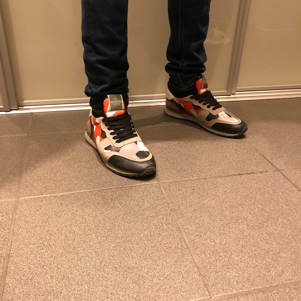 Säljer ett par snygga Valentino Rockrunner sneakers i ett camouflagemönster. Skorna har en mix av färger som orange, svart och grå med klassisk snörning. Perfekta för att ge din outfit en edgy touch. De är i bra skick. . Skor.