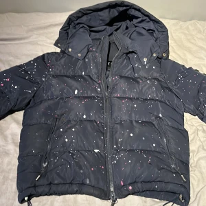 Mörk blå/svart pufferjacka från NVLTY - Säljer en svart/mörkblå pufferjacka från NVLTY med coolt färgstänk-mönster i vitt och rosa. Jackan har en mysig huva och dragkedja framtill. Perfekt för kalla dagar när du vill sticka ut lite extra. Den är i bra skick,jackan är mörkblå men ser ut som svart den är helt perfekt till vintern och kalla dagar inga problem med den alls, använd cirka 10 gånger för köpte ny inga problem med den skriv vid frågor eller funderingar