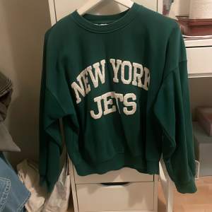 Säljer en grön sweatshirt med texten 'New York Jets' i vitt på framsidan. Tröjan har en avslappnad passform och långa ärmar, perfekt för en casual look. Den är i bra skick och passar perfekt för höst och vinter. Perfekt för sportfantasten eller som en cool vardagströja!