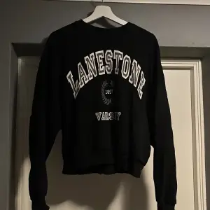 Säljer en svart sweatshirt från NLY Trend med trycket 'LANESTONE VARSITY' i vitt. Tröjan har en klassisk rund hals och långa ärmar, perfekt för en avslappnad stil. Den är i bra skick och passar perfekt för höst och vinter.